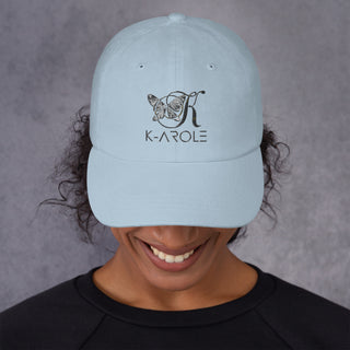 Light blue dad hat with K-AROLE logo embroidered, worn by smiling person with curly dark hair