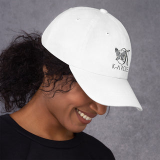 White K-AROLE brand baseball cap with logo, modeled by a smiling woman with curly hair against a grey background.