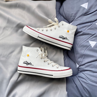 Trendy white high-top sneakers with delicate daisy flower embellishments, displayed against a gray and white striped background. The shoes feature a classic lace-up design and red accents, showcasing a stylish and casual aesthetic.